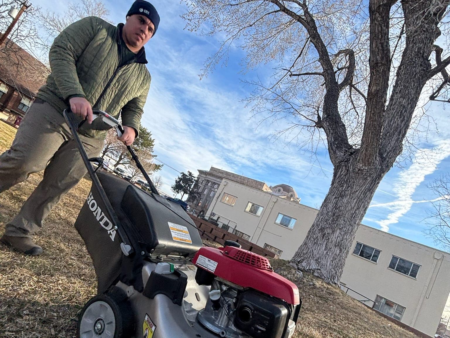 Crew member mowing a Pueblo lawn with a walk-behind mower under a bare cottonwood