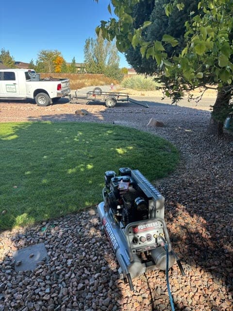 An air compressor set up on a rock bed beside a green lawn for a fall sprinkler blowout