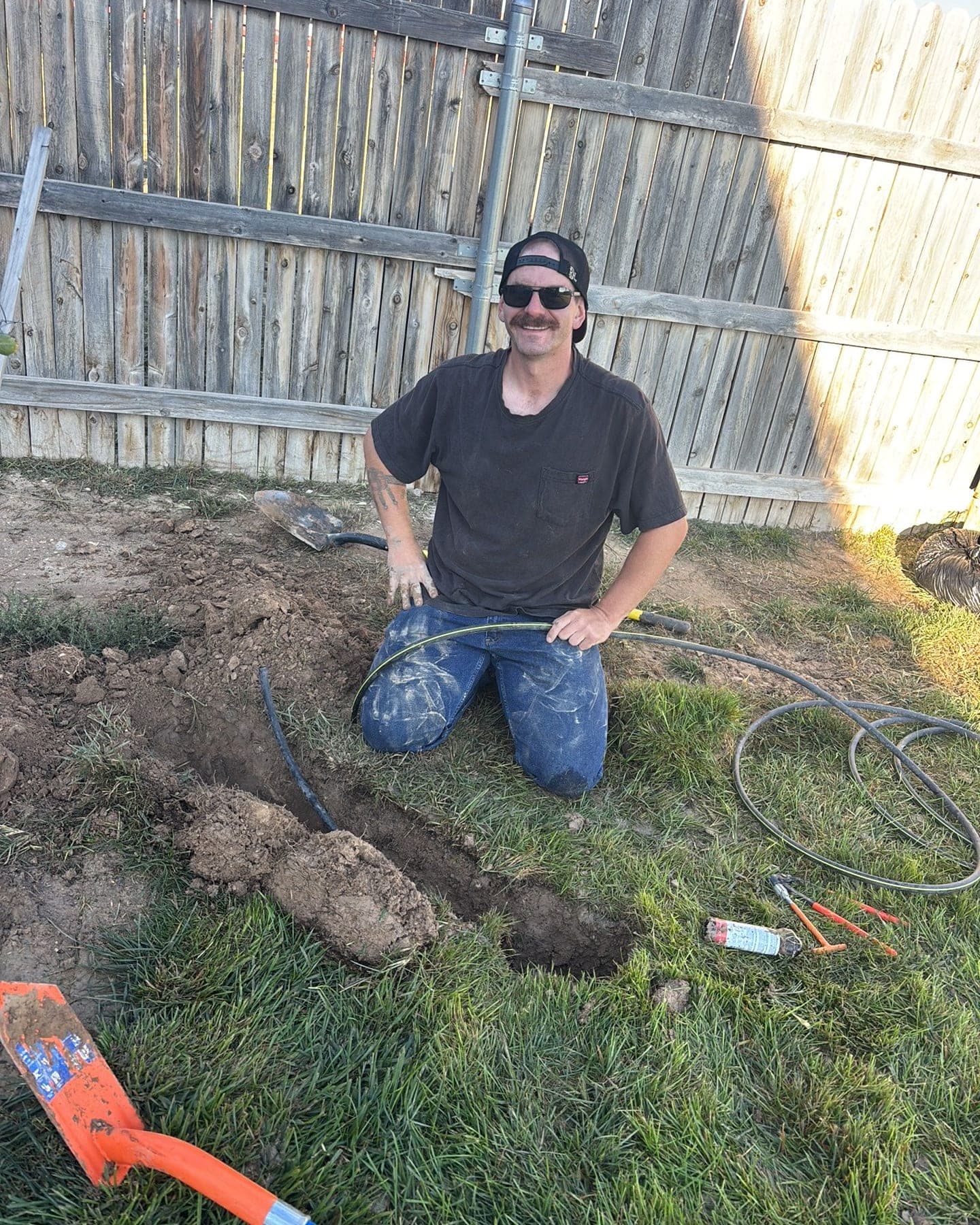Smiling crew member kneeling at a dug-out sprinkler line in a Pueblo backyard, shovel and fittings beside him
