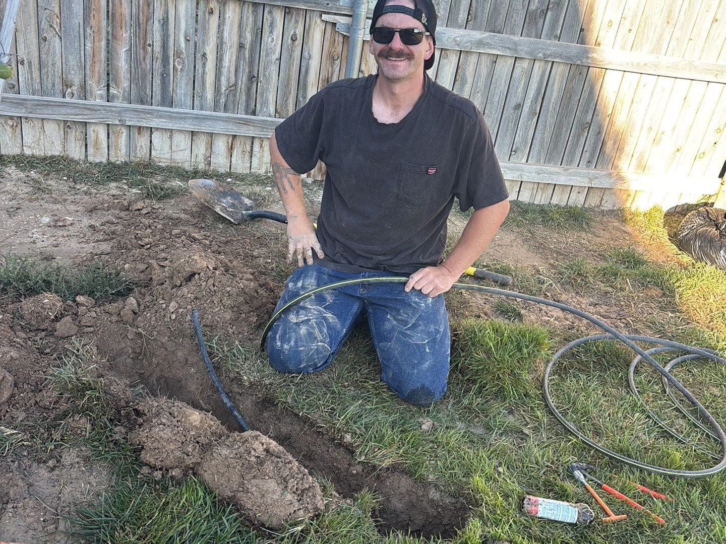 Smiling crew member kneeling at a dug-out sprinkler line in a Pueblo backyard, shovel and fittings beside him