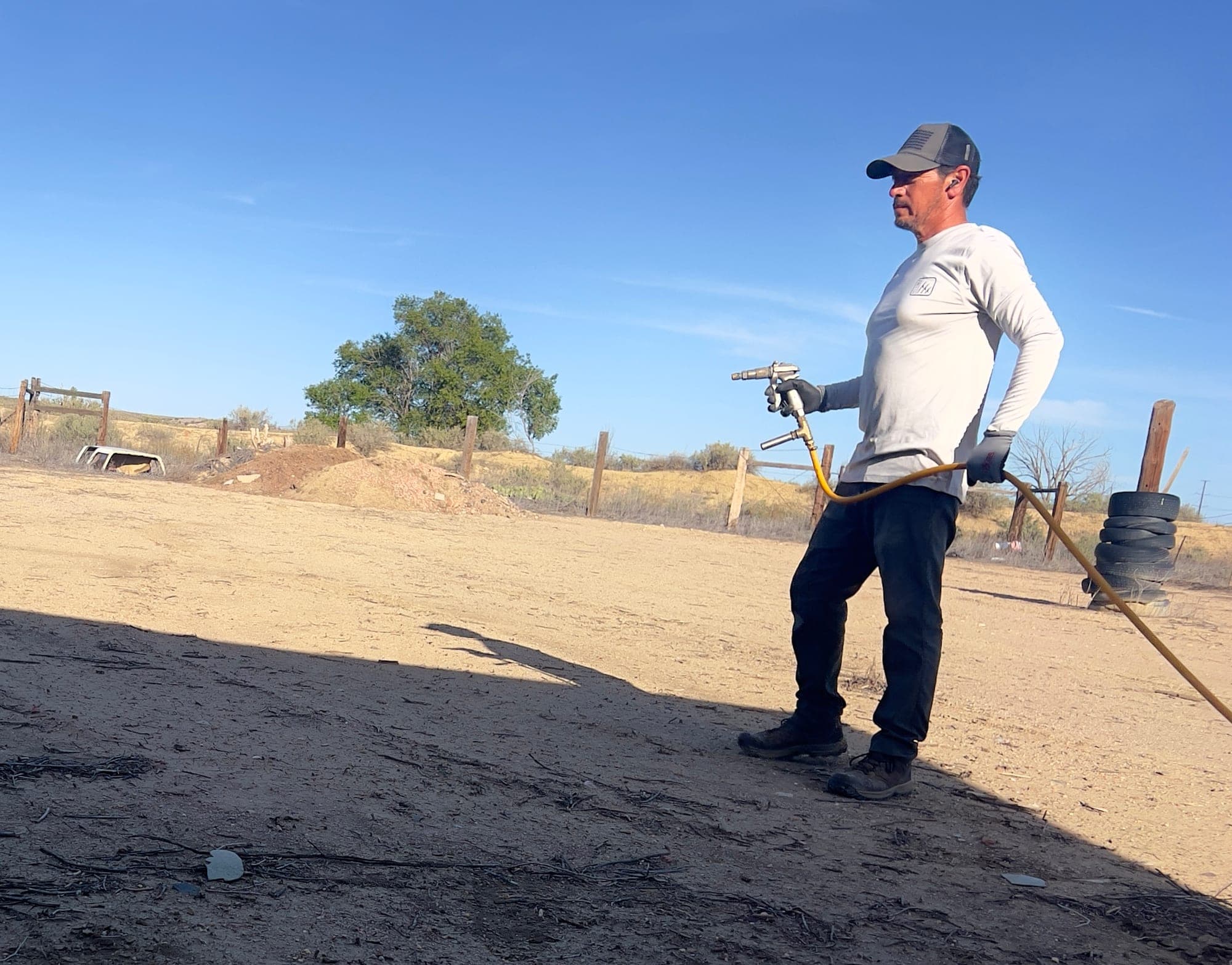 Crew member spraying weeds across a dirt lot in Pueblo with a hose-fed spray gun